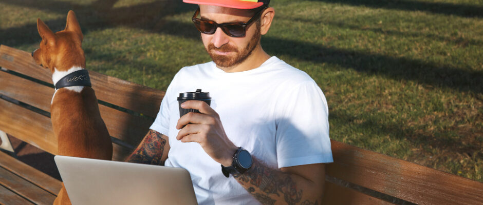 Young man with beard and tattoos wearing a plain white t-shirt drinking coffee and looking at his laptop and using rural internet while his brown and white dog sits next to him on a park bench.