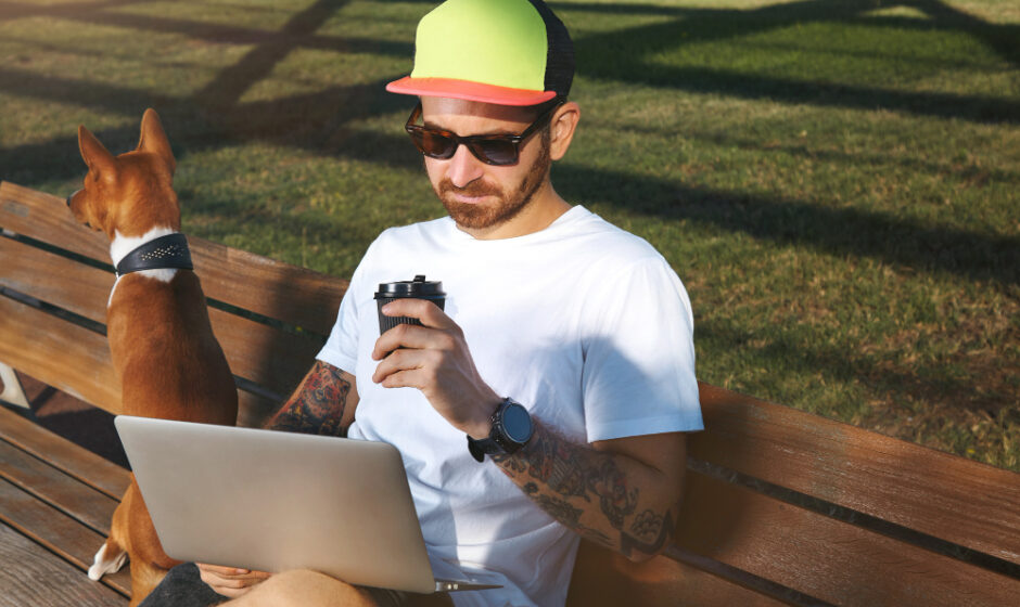 Young man with beard and tattoos wearing a plain white t-shirt drinking coffee and looking at his laptop and using rural internet while his brown and white dog sits next to him on a park bench.