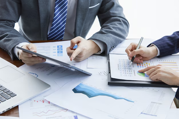 Business professionals analyzing financial reports and data, using a tablet and pen, with charts, graphs, and a laptop on a desk – collaborative financial planning and strategy meeting.