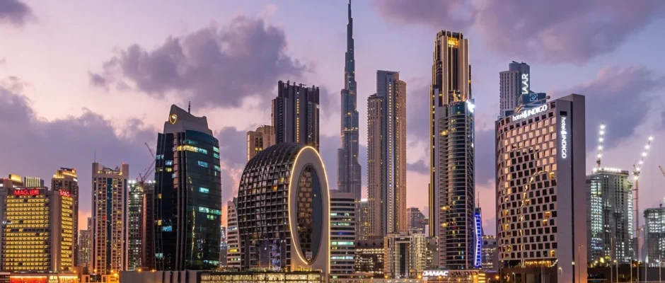 Skyline of Dubai at dusk, featuring modern high-rise buildings including the iconic Burj Khalifa in the center, uniquely designed skyscrapers like The Opus by Zaha Hadid with a central void, and illuminated waterfront properties reflecting on the calm water below.